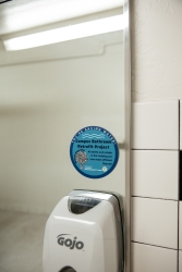 A blue circular sticker that is placed on a bathroom mirror on top of a soap dispenser. The sticker itself contains information for the Koffler Bathroom Retrofit Project. 