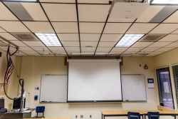 A classroom that has the lights on. A projector screen is down in front of a whiteboard.