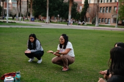 Two people squatting down next to each other on a grass field while the person on the right talks to a group of people in a circle..