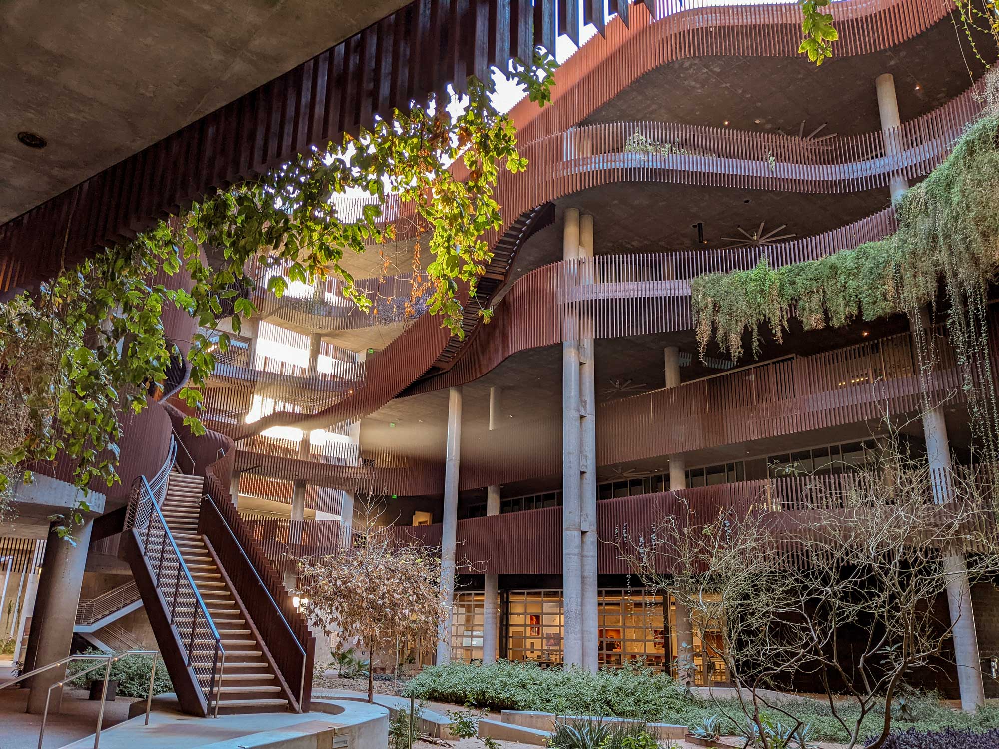 The Environment & Natural Resources 2 (ENR2) building at the University of Arizona serves as a flagship for sustainable design, featuring a striking multi-level structure with curved walkways, exposed concrete columns, and rust-colored metal accents, surrounded by lush greenery and cascading plants that embody its commitment to environmental responsibility.