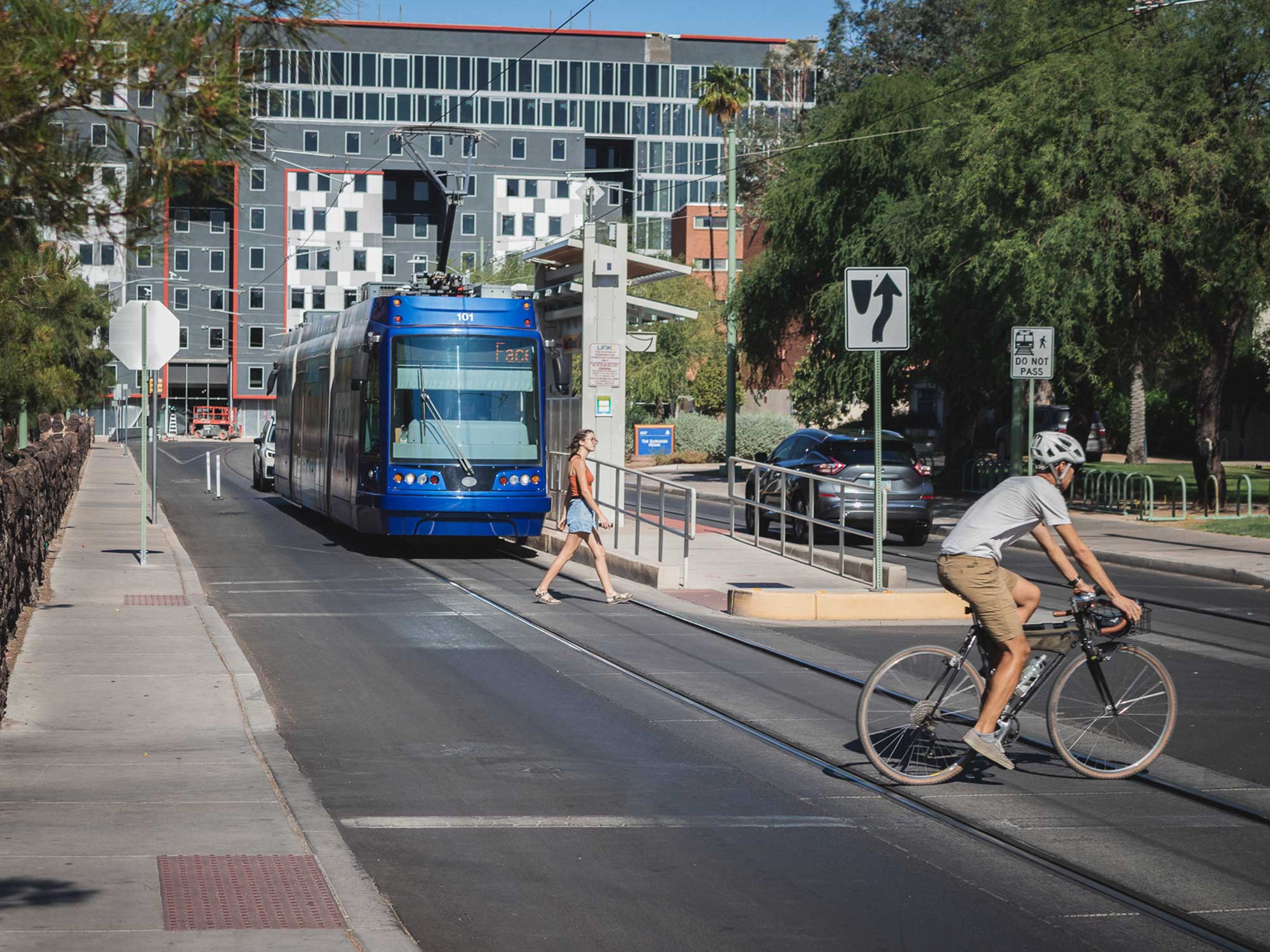 A modern blue streetcar moves along tracks in an urban setting as a woman crosses at a designated pedestrian crossing and a cyclist rides past, with a multi-story building, green trees, traffic signs, and public transportation infrastructure in the background.