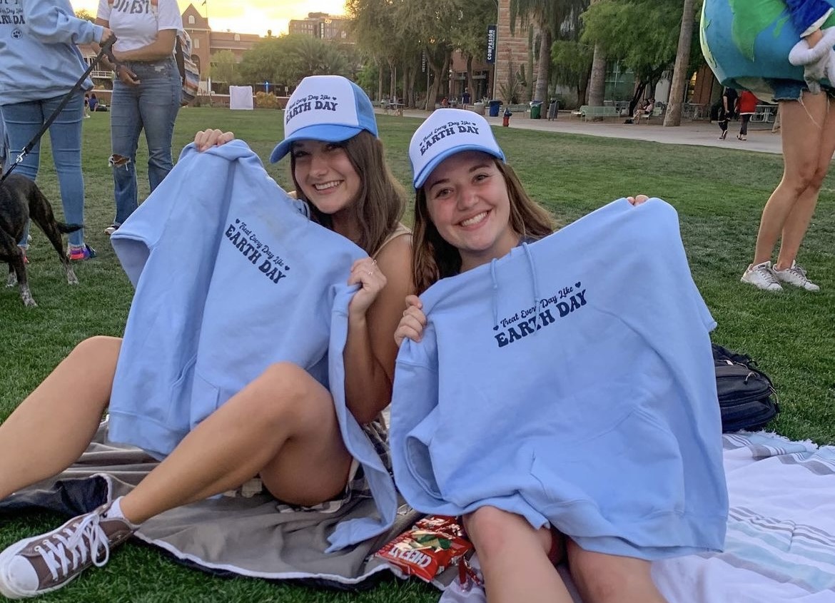 Two students sit on the ground holding up shirts that read “Earth Day”.