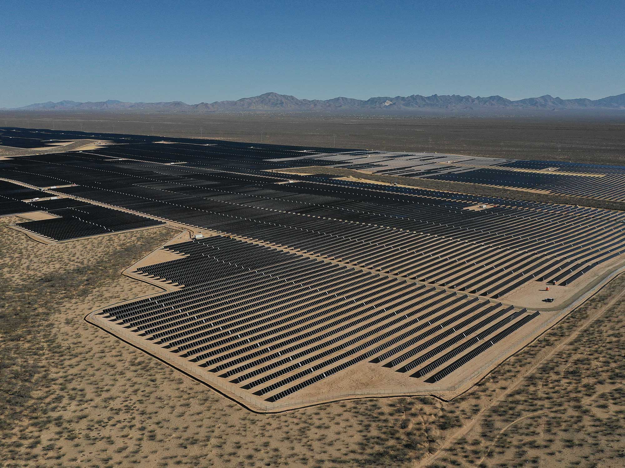 An expansive solar farm stretches across the arid desert landscape, with countless rows of solar panels aligned in geometric patterns under a clear blue sky, backed by distant mountain ranges.