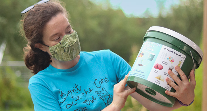 Woman reading the label of a Bucket Program compost bucket, wearing a cloth face mask