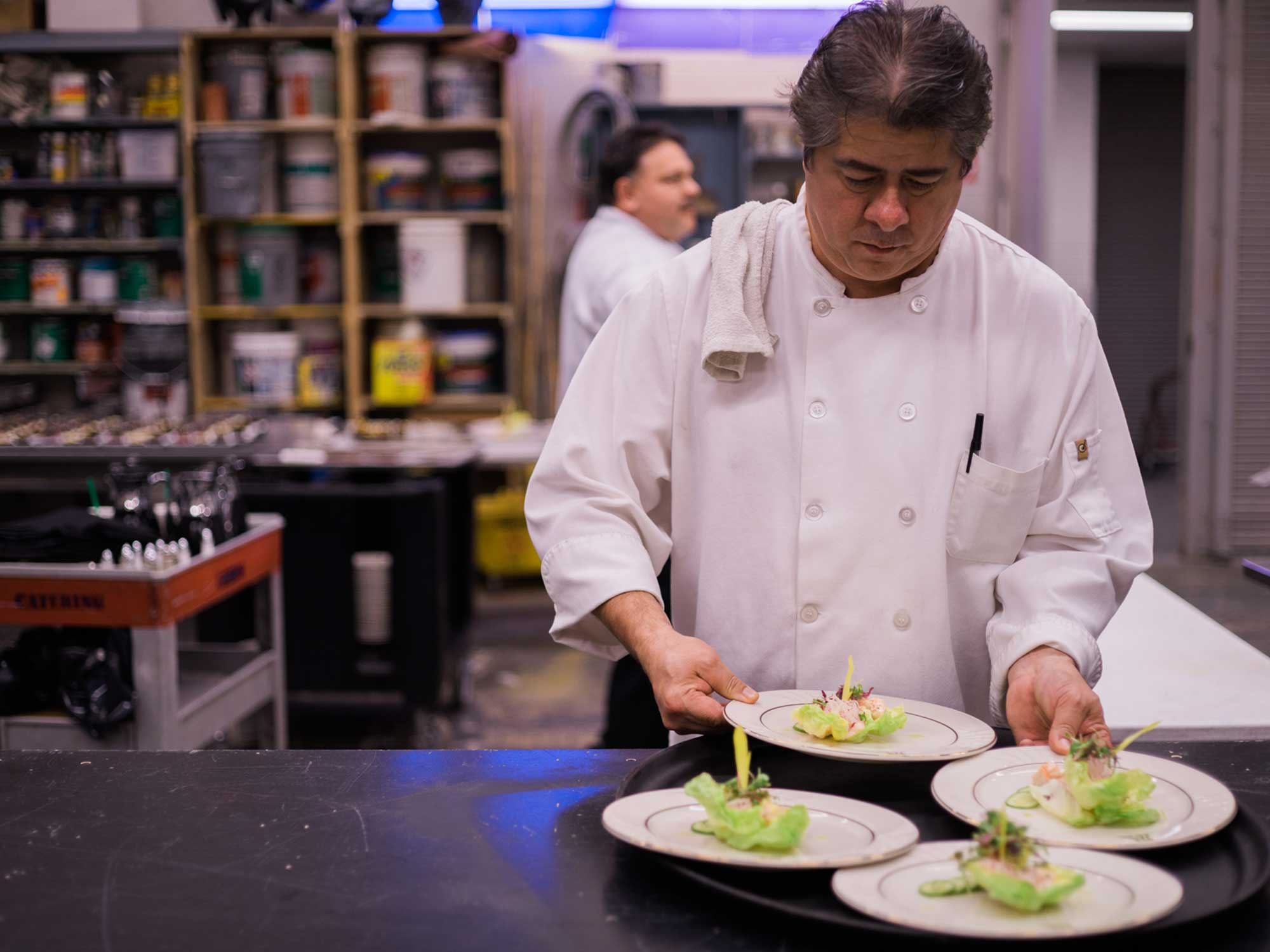 A chef in a white uniform carefully arranges plates of beautifully presented plant-based dishes, featuring fresh greens and garnishes, in a professional kitchen setting with another chef and stocked shelves in the background.