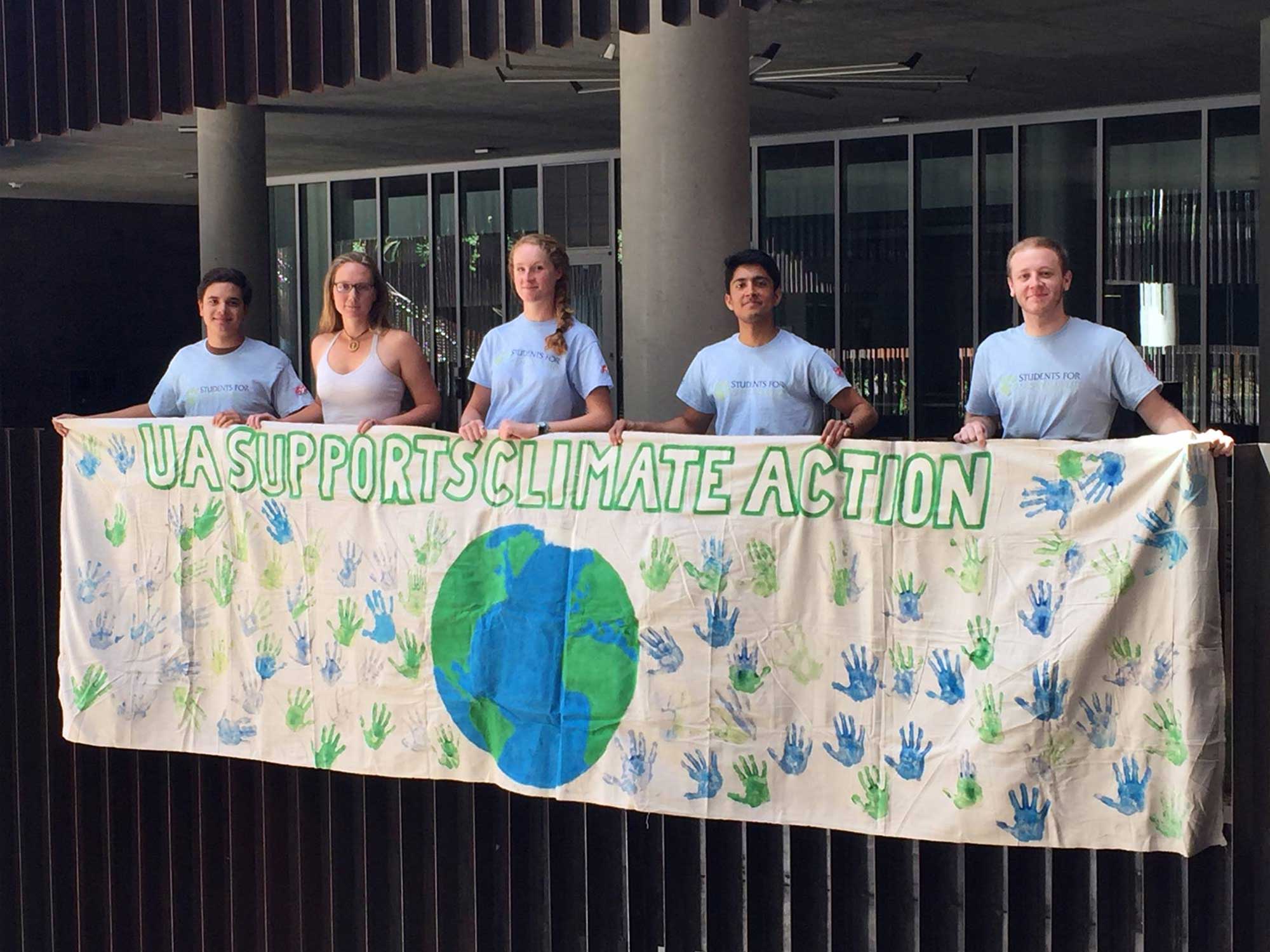 A group of students stands in front of the Environment & Natural Resources 2 (ENR2) building at the University of Arizona, holding a handmade banner that reads 'UA Supports Climate Action, decorated with blue and green handprints surrounding a painted Earth.
