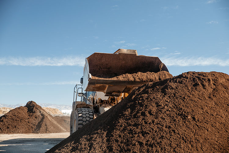 A large tractor pours additional compost on top of an 8 foot tall pile. Another pile of compost rises in the background.