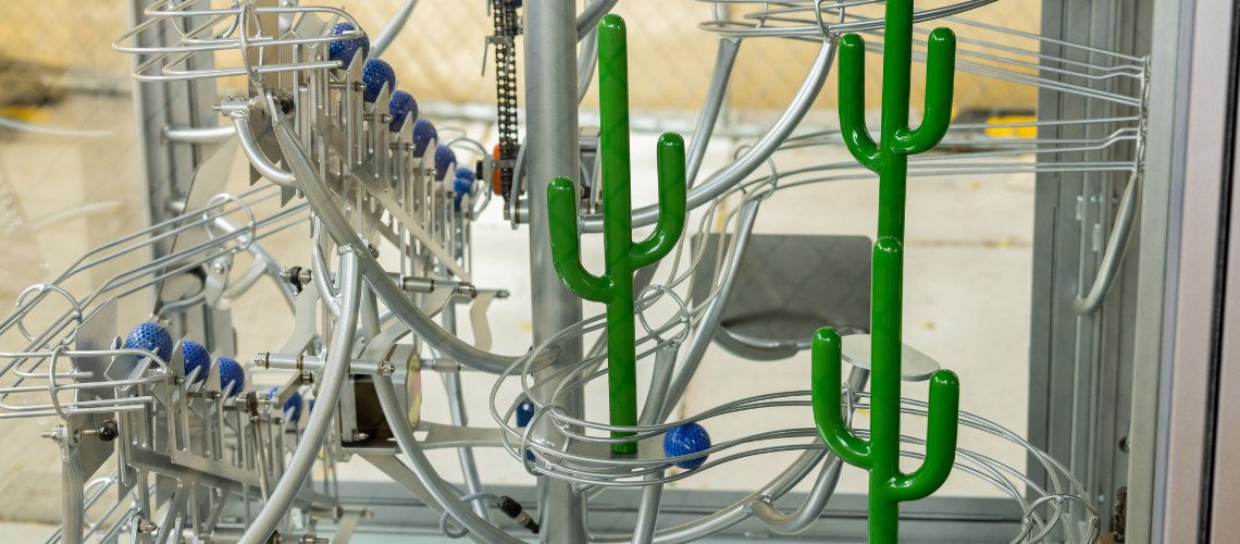 Blue golf balls roll down track behind miniature saguaros in a ball machine display.