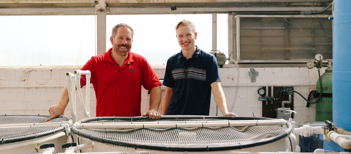 Project Manager Benjamin Hunt and Advisor Dr. Recsetar standing in front of a fish tank inside a greenhouse