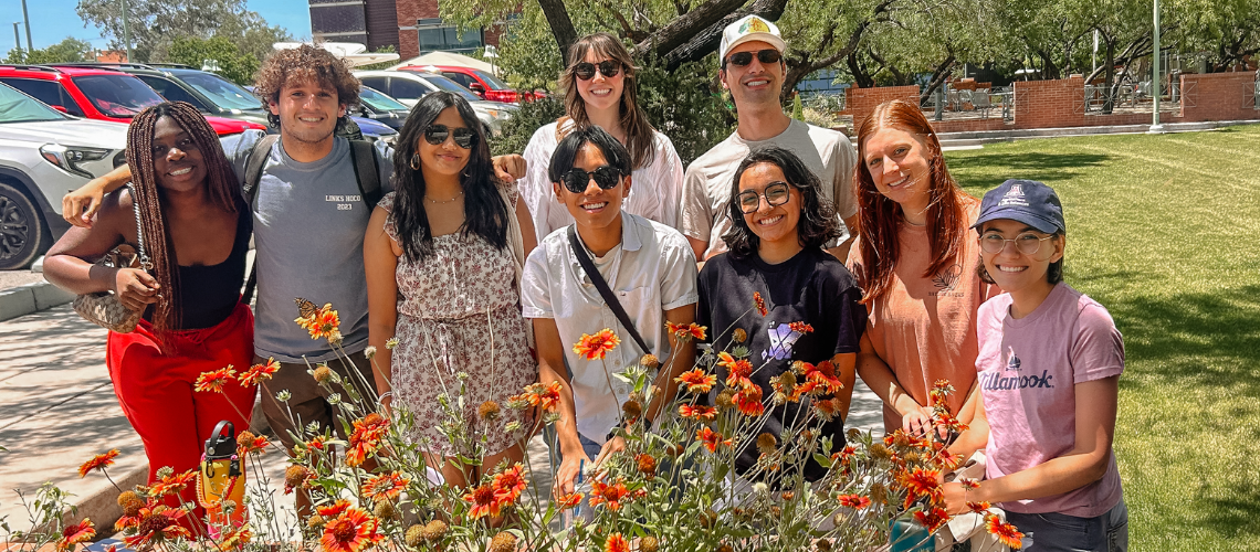 A group of people standing next to each other and smiling behind a flower bush.