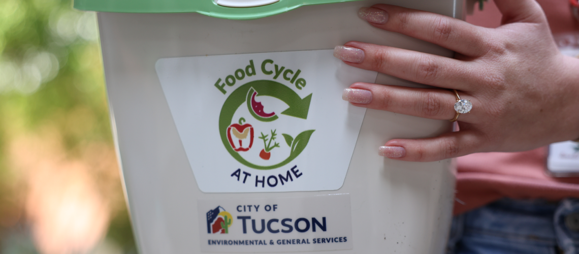 A very large diamond engagement ring sparkles on a hand holding a bucket for collecting food scraps with Food Cycle at Home and City of Tucson labels on it
