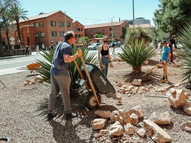 A group of people standing in a dirt area holding various types of gardening tools. One person is holding a wheelbarrow.
