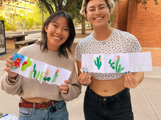 Two people smiling and holding drawings that are folded in a pamphlet style that include trees and plants.
