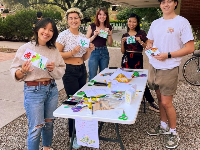 A group of people standing around a table, smiling and holding drawings on paper that is folded in a pamphlet style.