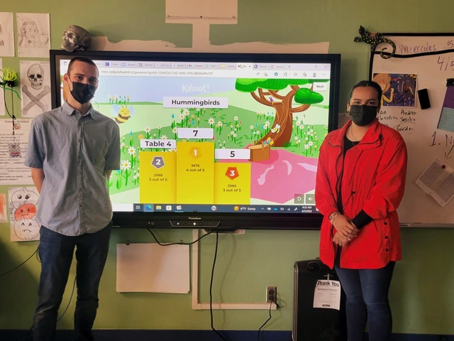 2 teachers in a classroom standing in front of a tv screen.