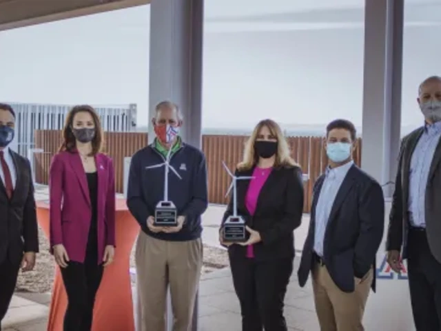 A group of six professionally dressed individuals stand side by side outdoors under a covered structure, all wearing face masks. Two individuals in the center are holding small wind turbine-shaped awards.
