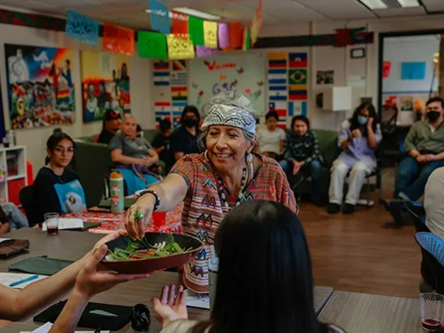 In a vibrant, communal space decorated with papel picado banners, cultural artwork, and international flags, a woman in a colorful woven top and patterned headscarf smiles warmly as she shares a bowl of fresh greens with a participant, while attendees engage in conversation and learning.