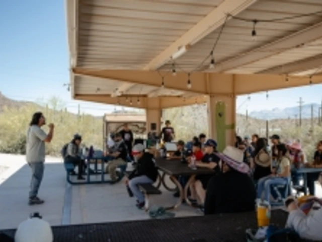 Students listen intently to an educator who is facing them and providing a lesson underneath an outside ramada.