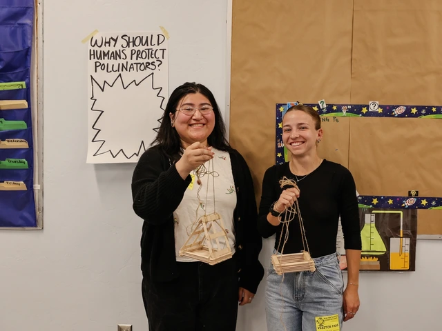 Two people standing next to each other, smiling, and holding up structures made out of wooden popsicle sticks.