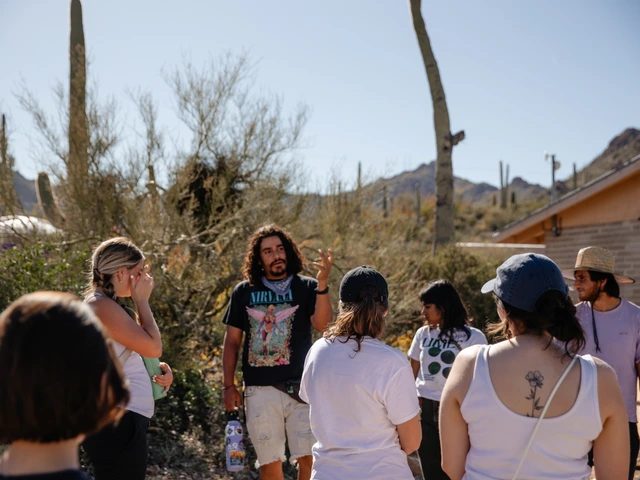 A group of people standing outside in a desert area, looking at a person talk to them.