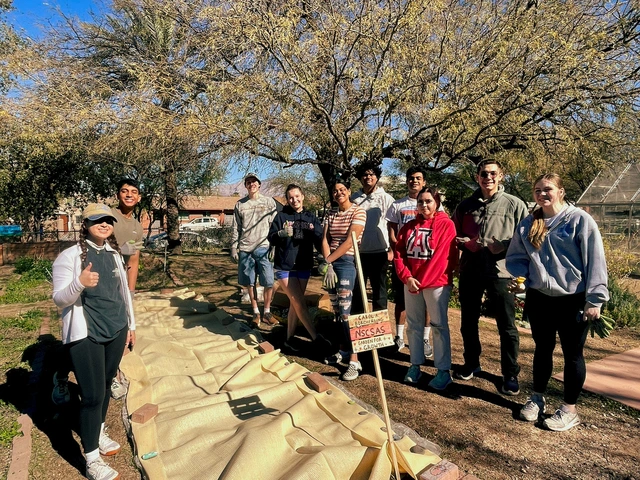 A group of people posing for a photo next to a garden plot that is covered with a sun protecting tarp.