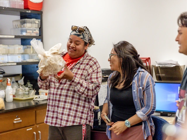 Three people are standing next to each other while all looking at the person to the left. The person on the left is holding a bag with a large white mushroom inside of it.