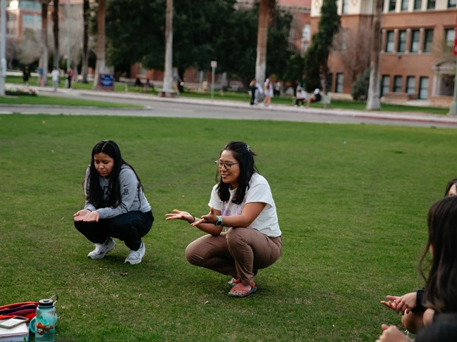 Two people squatting down next to each other on a grass field while the person on the right talks to a group of people in a circle..