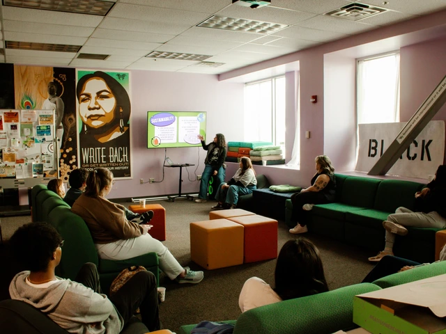 A group of people sitting around a common area looking at a tv. A person next to the tv has their hand raised to point at the screen.
