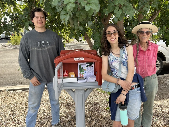 Three people stand under a tree next to a small seed library