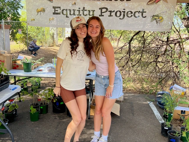 Two people hugging and smiling in front of a hanging sign that reads "Pollinator Equity Project".