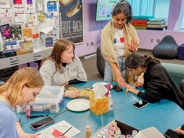 A group of people sitting at a table painting. One person is standing while getting their nails painted by another person sitting next to them. 