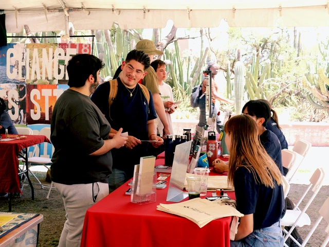 People standing next to a table covered in outreach material. They are holding pamphlets and talking to the people sitting behind the table.