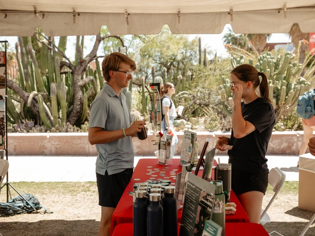 Two people talking to each other with a red table in between them. One person is holding a jar filled with compost.