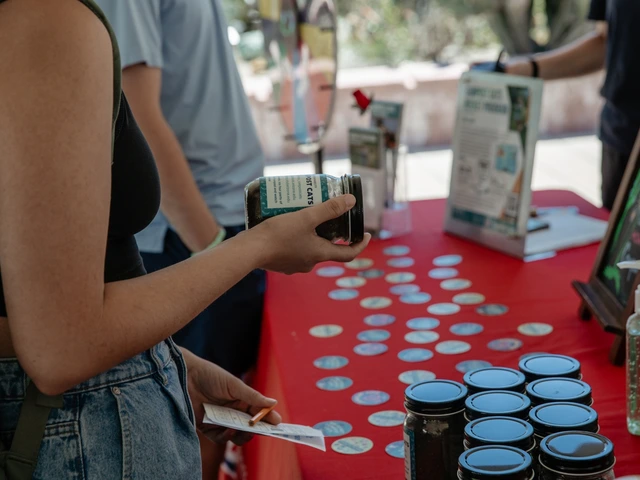A table covered in red cloth that is filled with stickers, jars filled with compost, and informational cards. A person is standing near the table holding a compost jar.