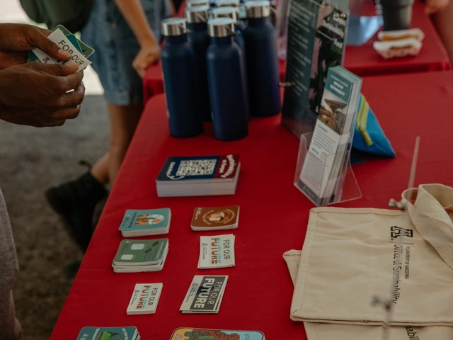 A table covered in red cloth that is filled with stickers, water bottles, and tote bags. A person's hand is shown holding stickers.