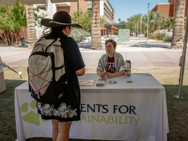 A person talking to another person that is sitting behind a table covered in white cloth. "Students for Sustainability" is written on the white cloth.