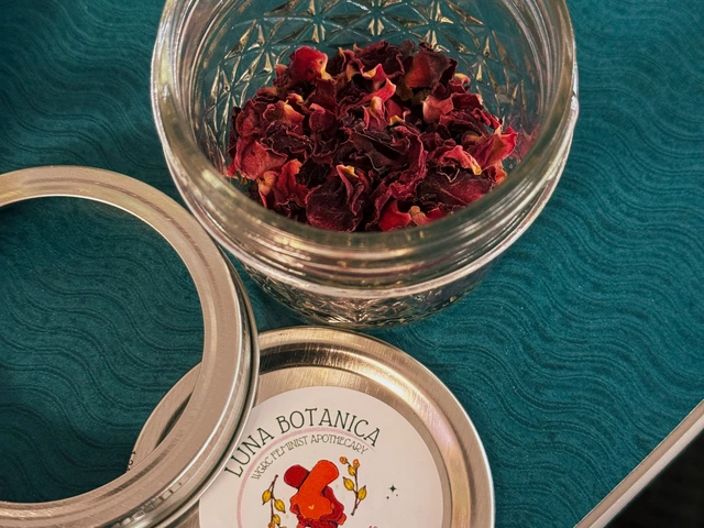 A small glass jar has rose petals inside it. The lid of the jar is laid out next to the glass jar with a circular sticker on top of the lid.