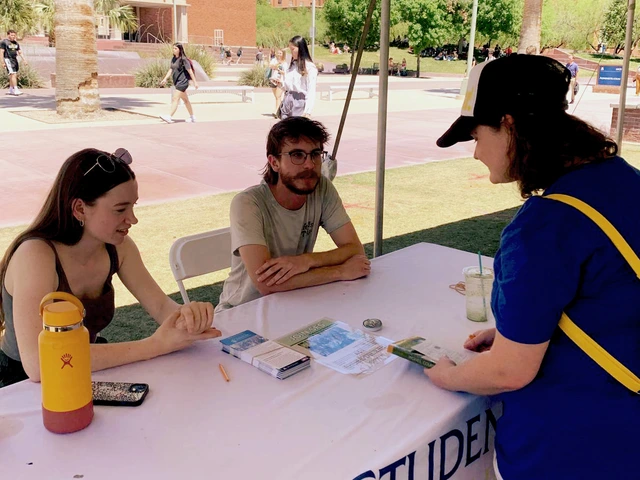 A person talking to two other people who are sitting behind a table covered in white cloth.