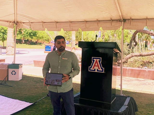 A person stands in front of a podium talking to a group of people under a white tent.