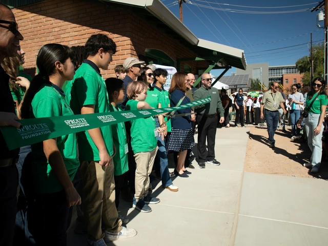 A child stands in a line of people wearing green, poised to cut a green ribbon saying Sprouts Foundation with a pair of large scissors.