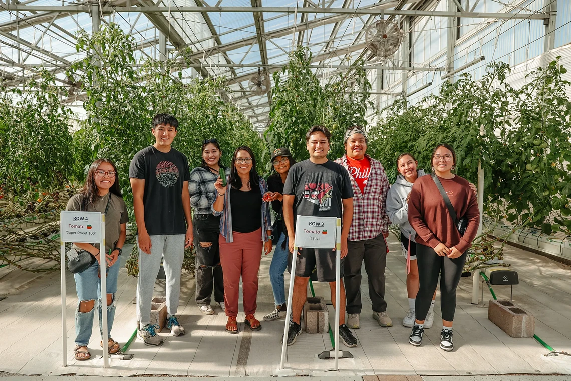 A group of people stand inside a greenhouse with rows of tomato plants behind them. Two signs are visible: one reads 'ROW 4 Tomato 🍅 Super Sweet 100,' and the other reads 'ROW 3 Tomato 🍅 Estiva'.