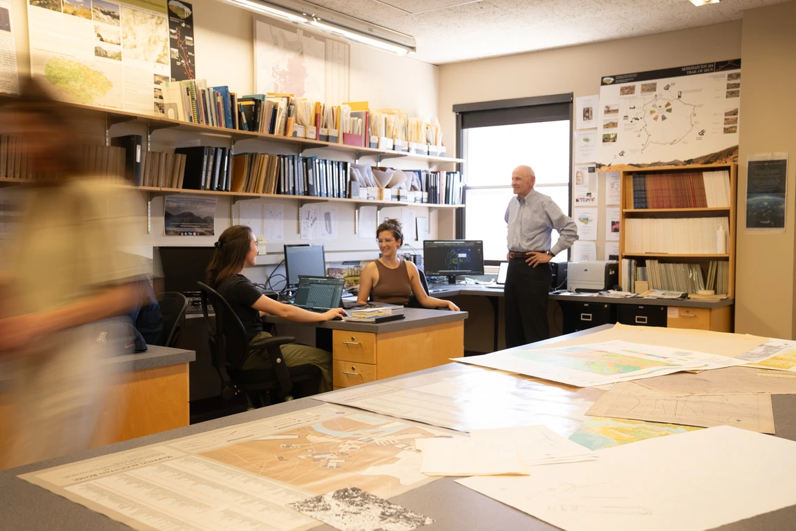 A group of three people sit and talk together in a room full of maps.