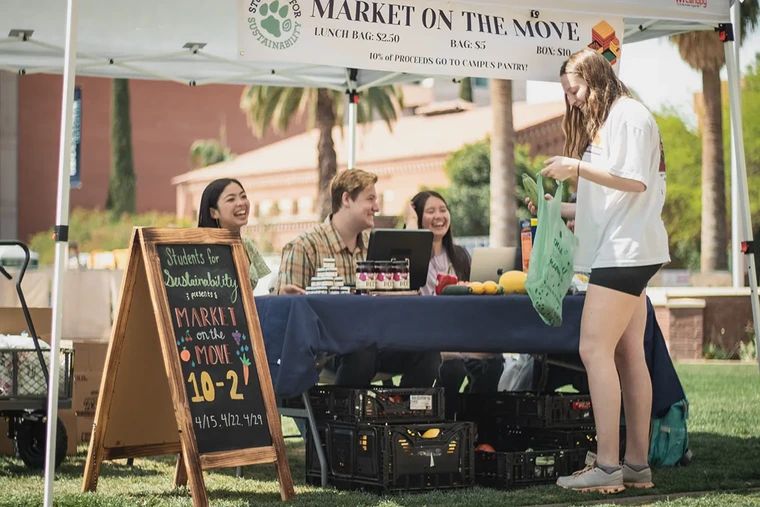 An outdoor market stall under a canopy with a sign reading MARKET ON THE MOVE operated by Students for Sustainability.