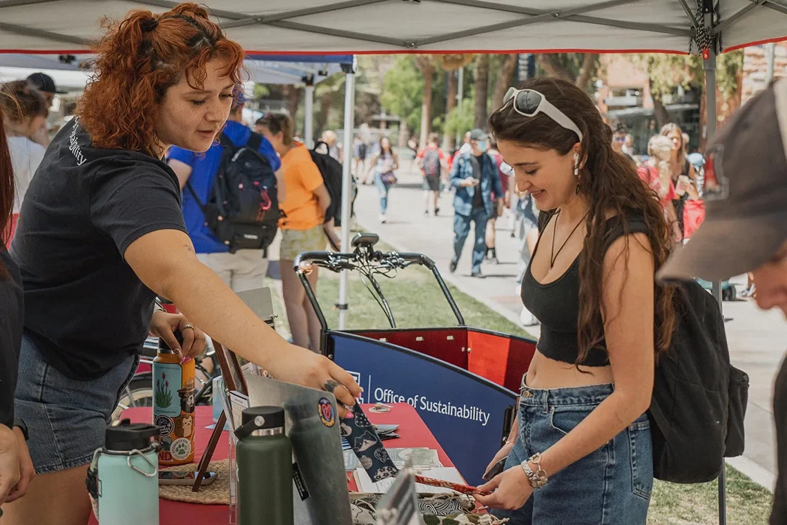 An outdoor event booth under a canopy tent labeled 'Office of Sustainability.' The table displays water bottles, pamphlets, and promotional items. People interact at the booth, with one person handing something to another. Other booths and attendees are visible in the background.