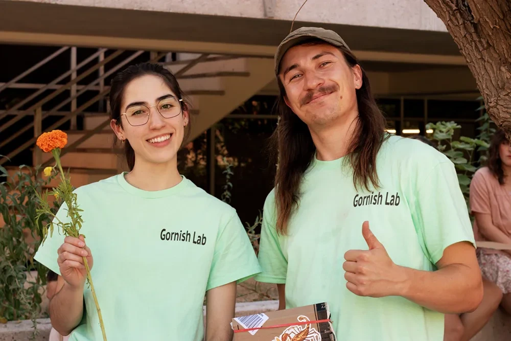 Two people smiling. One is holding a orange flower and the other is holding a thumbs up.
