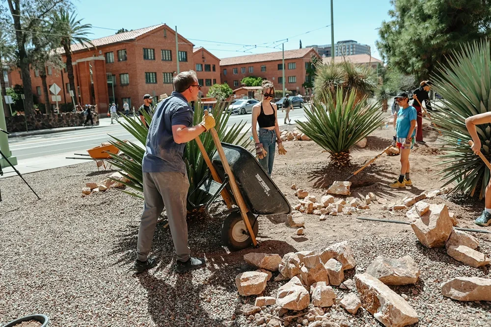 A group of people standing in a dirt area holding various types of gardening tools. One person is holding a wheelbarrow.