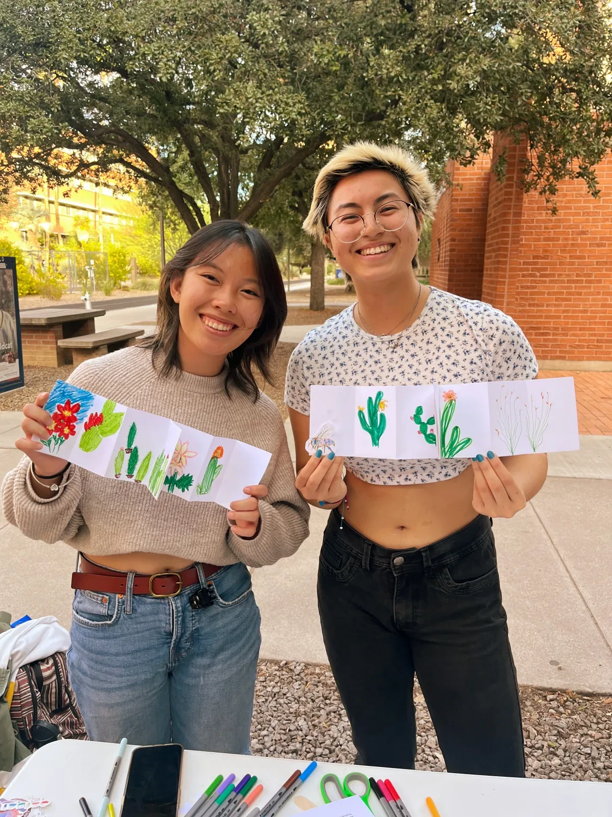 Two people smiling and holding drawings that are folded in a pamphlet style that include trees and plants.