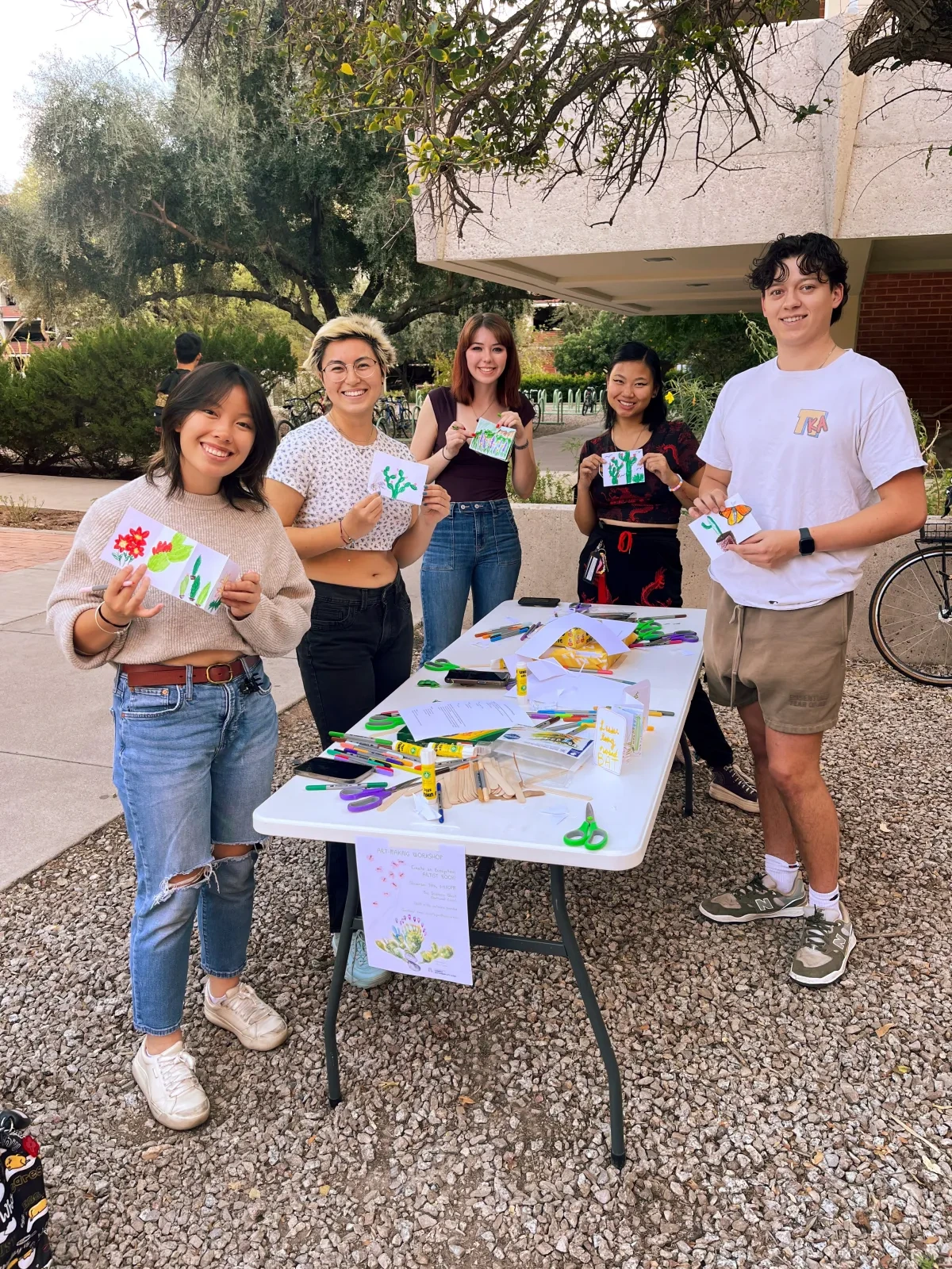 A group of people standing around a table, smiling and holding drawings on paper that is folded in a pamphlet style.