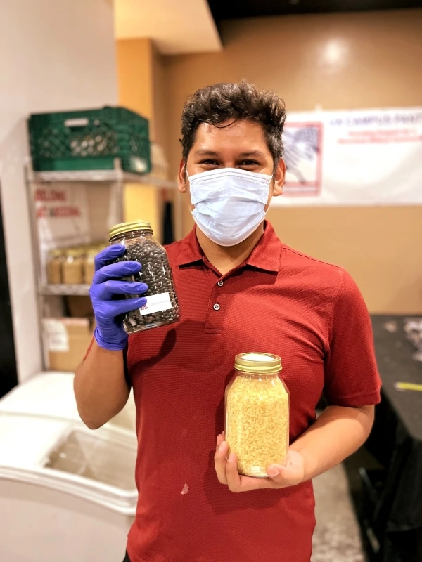 One individual in a mask holds up two glass mason jars that have dry goods in them for distributing food
