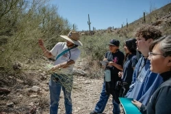 An educator is separating branches and explaining something to a group of students who are standing in a small wash.